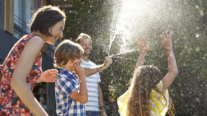 A father sprays a hose on a sunny day while his children run through the water and his wife smiles. 